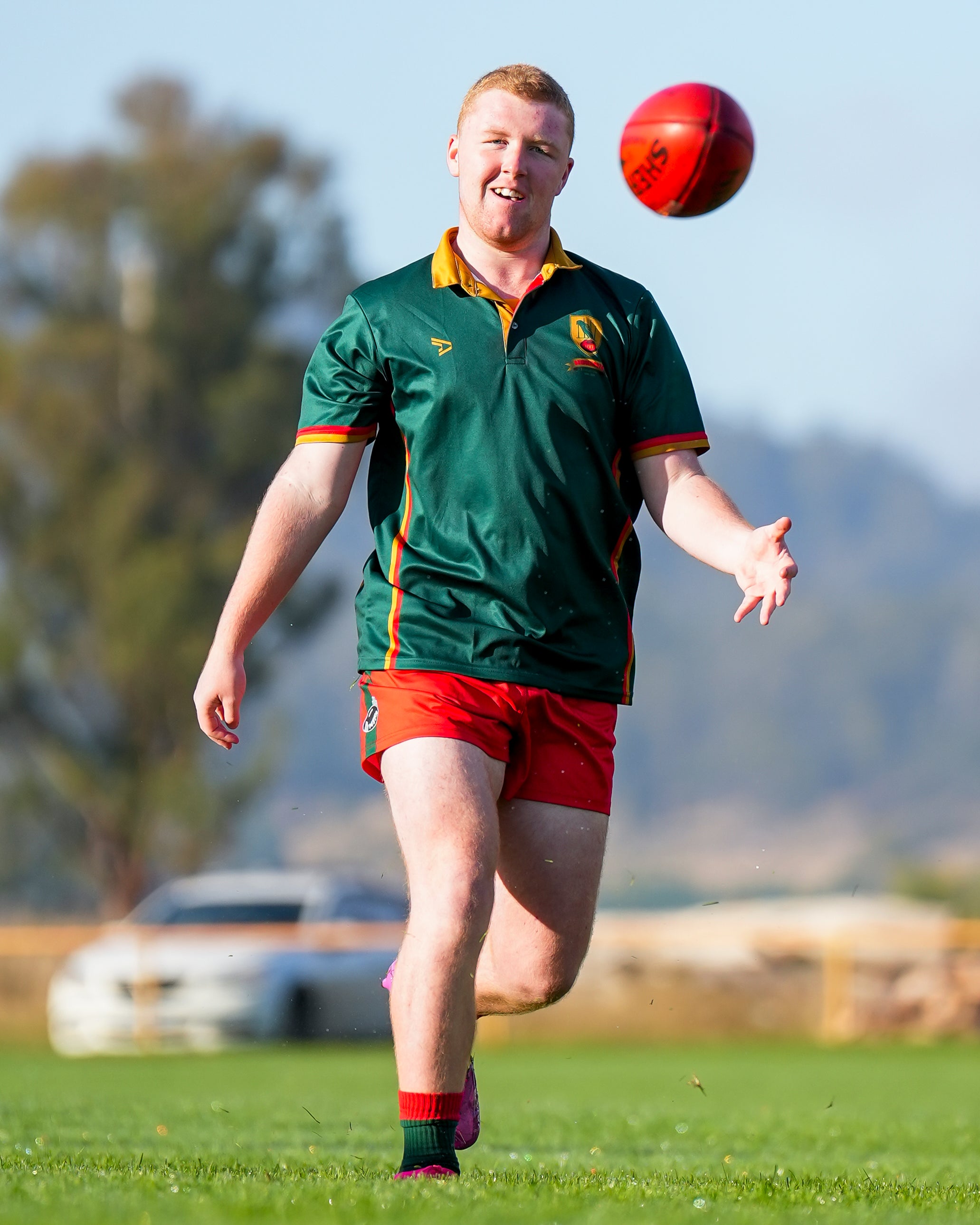 Bridgnorth player in uniform kicking a red ball on a grassy field with trees and mountains in the background.
