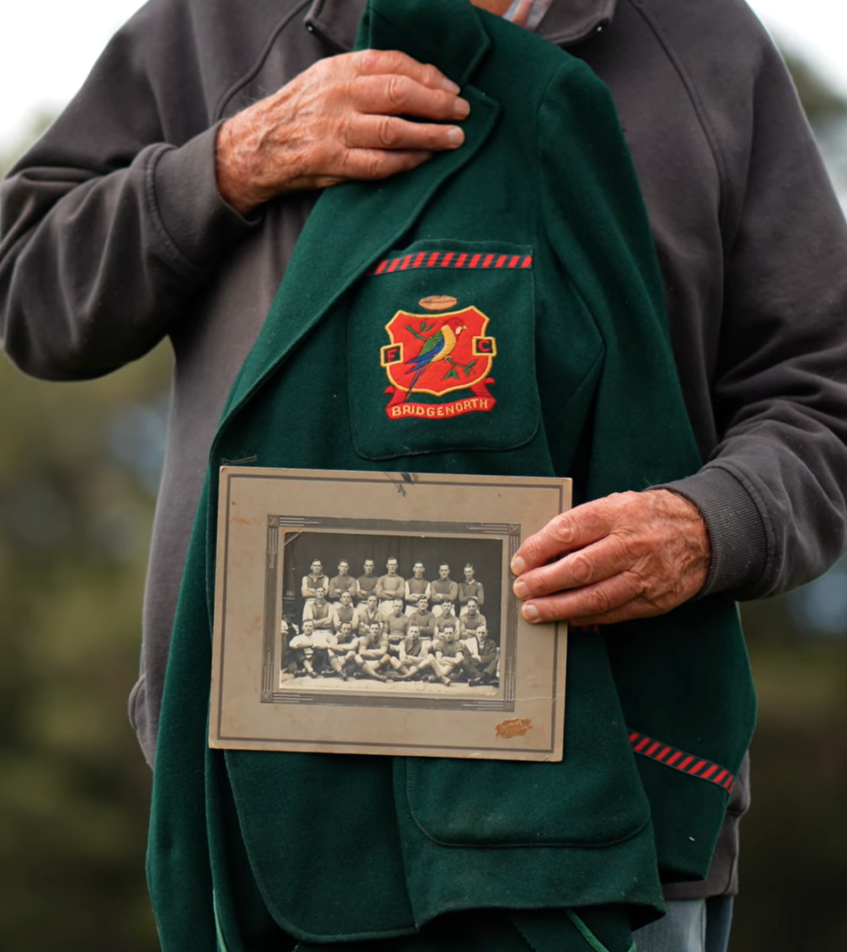 Person holding a framed black and white photo of the premiership team of 1929 inside a green jacket with a logo.