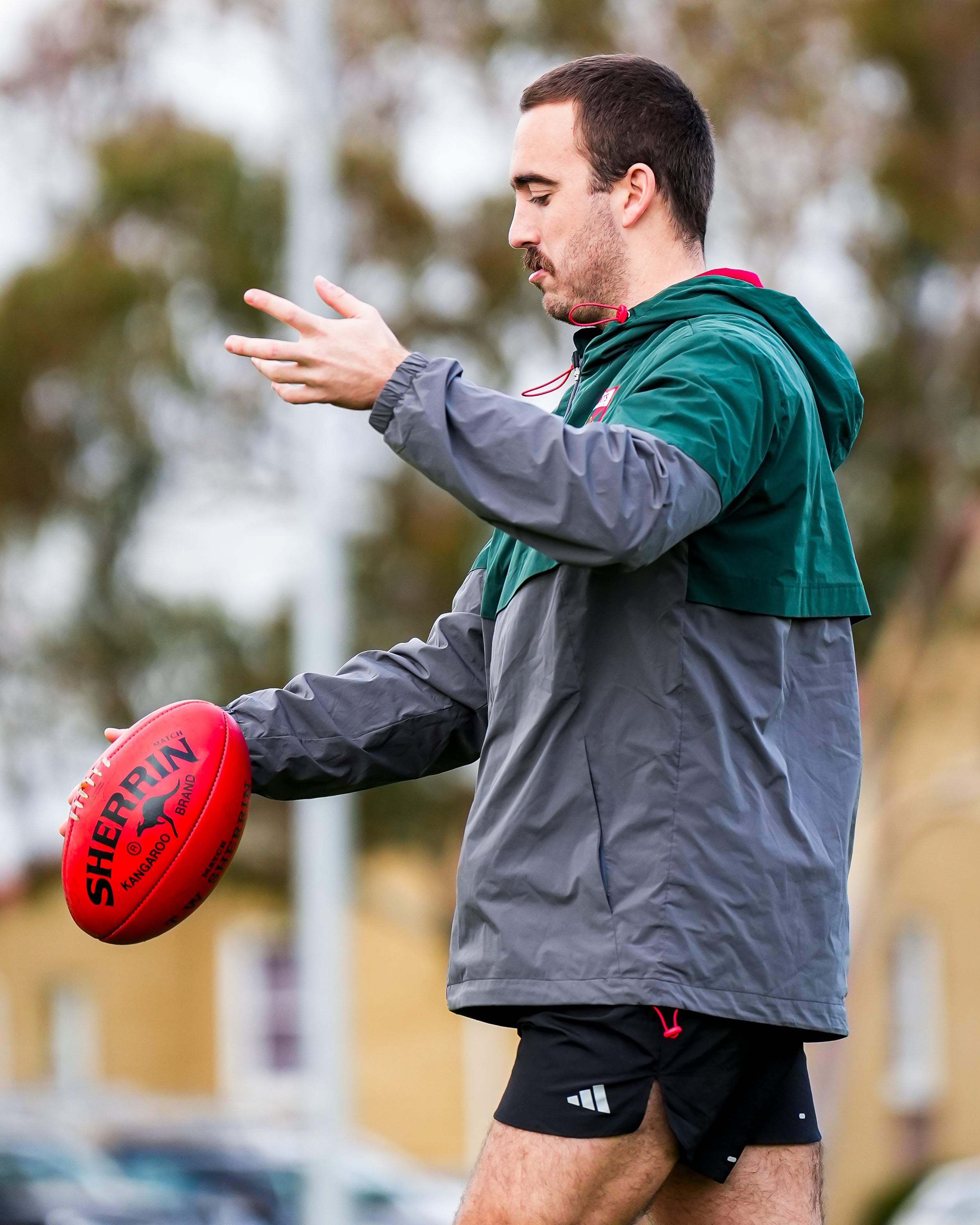 Man holding a red AFL ball outdoors with trees and buildings in the background